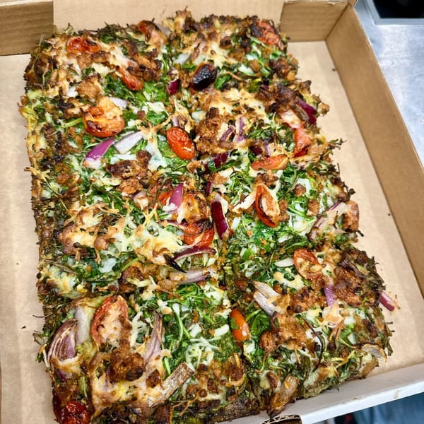 A rectangular vegan pizza with green vegetables, red onions, cherry tomatoes, and vegan cheese in a box.