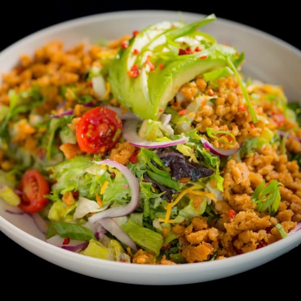 A vibrant vegan salad with lettuce, tomatoes, red onions, avocado, and plant-based crumbles in a white bowl.