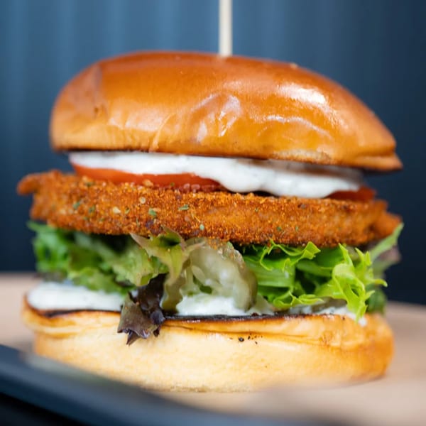 Close-up of a vegan crispy chicken sandwich with lettuce, tomato, and white sauce on a bun.