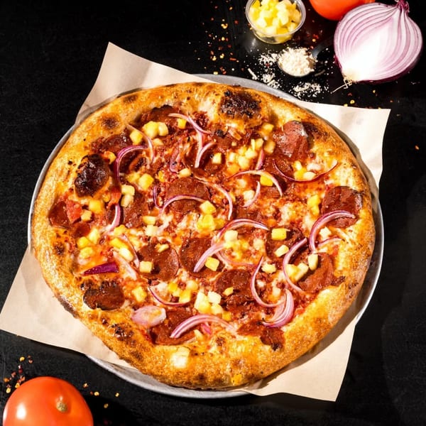 Vegan pizza with pepperoni, pineapple, and red onion, on a dark surface with ingredients.