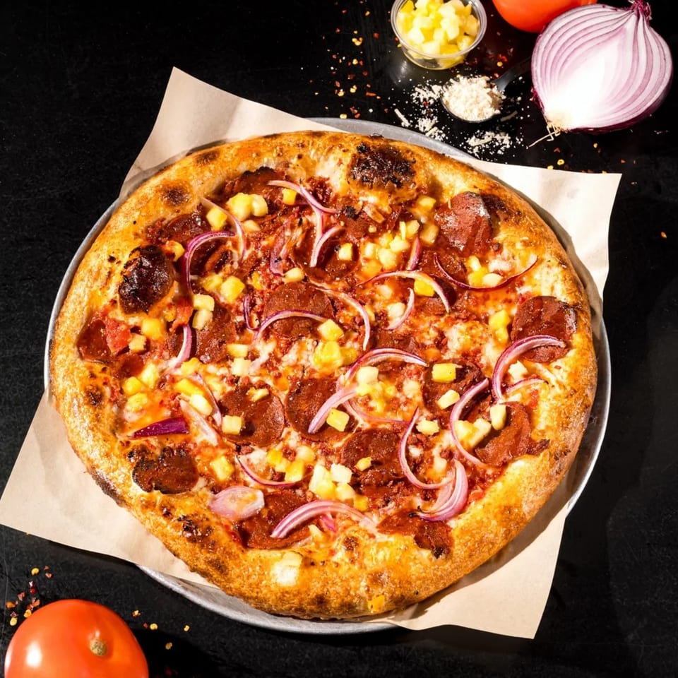 Vegan pizza with pepperoni, pineapple, and red onion, on a dark surface with ingredients.