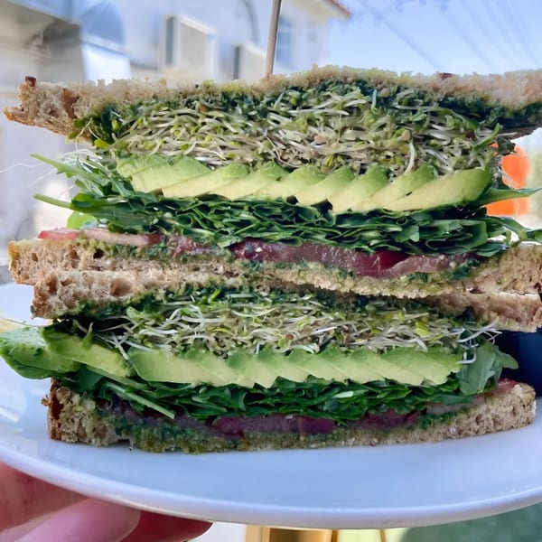 Stacked vegan sandwich with sprouts, avocado, arugula, and tomato on whole wheat bread.